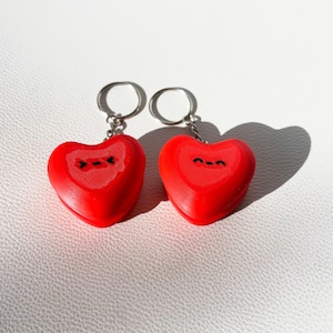 May include: Two red heart-shaped keychains with black smiling faces. Each keychain has a silver metal ring attached to the top. The keychains are on a white surface, and a shadow of the keychains is visible.