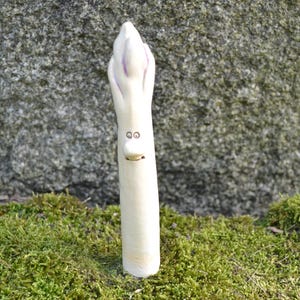 May include: White ceramic sculpture of an asparagus with a pink top and a painted face. The sculpture is standing on a bed of green moss.