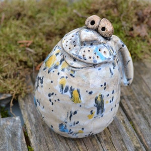 Può includere: Una statuetta in ceramica di una creatura fantasiosa con occhi grandi, una glassa bianca, blu e gialla macchiata e un piccolo manico. La statuetta ha una superficie testurizzata e un'espressione giocosa.