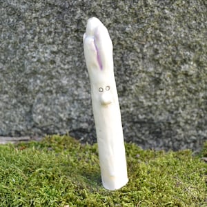 Könnte beinhalten: Weiße Keramikskulptur eines Spargels mit einer violetten Spitze und einem bemalten Gesicht mit zwei Augen und einer Nase.