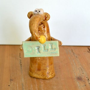 May include: A brown ceramic figurine of a creature with large eyes and a long nose holding a sign that says "DILL".