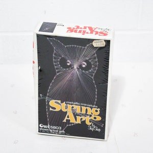 Puede incluir: Un kit de arte de hilo en miniatura metálico con un diseño de búho. El kit incluye todos los materiales necesarios para crear el búho, incluyendo hilo, clavos y una base de madera. El kit está empaquetado en una caja con una imagen en blanco y negro del búho terminado en la parte delantera. La caja también incluye el texto "String Art" y "Owl 5803 shown actual size."