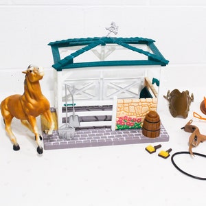 Op de afbeelding: Een plastic speelgoedpaardenstal met een bruin paardenfiguurtje, een groen dak en een wit hek. De set bevat een verscheidenheid aan accessoires, zoals een zadel, hoofdstel en borstels.