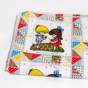 May include: A white fabric item with a colorful patchwork border. The center features a vintage illustration of two children, one offering a flower. Text includes "joan walsh anglund" and "a friend is someone who likes you."
