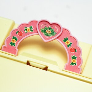 May include: A pink plastic handle with a heart-shaped cutout and a floral design. The handle has a scalloped edge and is decorated with small fruit and flower motifs.