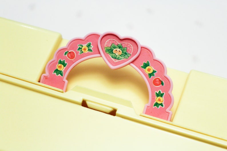 May include: A pink plastic handle with a heart-shaped cutout and a floral design. The handle has a scalloped edge and is decorated with small fruit and flower motifs.