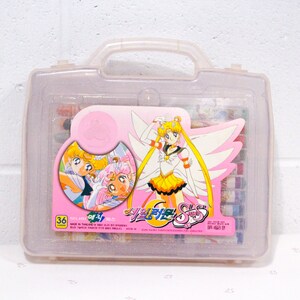 Puede incluir: Una caja de transporte de plástico rosa con una tapa transparente. La caja tiene un asa y está decorada con un personaje de Sailor Moon y el texto "세일러문 S" en coreano. La caja contiene 36 lápices de colores.