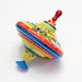 May include: A colorful spinning top toy with a yellow car design and a red handle. The top is made of metal and has a blue, yellow, red, and green striped design.