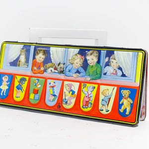 Puede incluir: Caja de juguetes de hojalata vintage con un diseño colorido. La caja presenta ilustraciones de niños mirando por las ventanas y varios personajes de dibujos animados. La caja tiene un marco negro con detalles en rojo y amarillo.