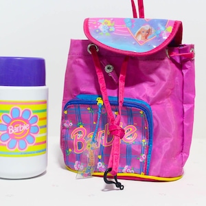 Puede incluir: Una mochila rosa de Barbie y un termo blanco con tapa morada. La mochila tiene un bolsillo frontal con el logo de Barbie y una solapa con una imagen de Barbie. El termo tiene un diseño floral con el logo de Barbie.