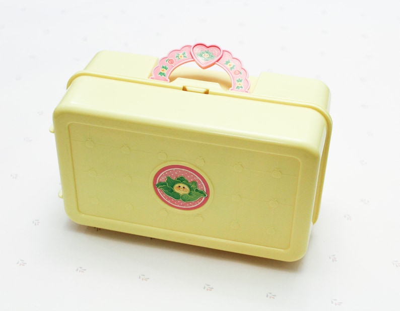 May include: A yellow plastic lunchbox with a pink and green floral design. The lunchbox has a handle shaped like a heart with a flower design.