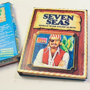 Puede incluir: Un álbum de sellos vintage Seven Seas World-Wide Stamp Album con una ilustración colorida de un pirata con un parche en el ojo y un loro en su hombro. La portada del álbum es marrón con ribetes dorados y el título está en letras blancas en negrita.