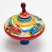 May include: A colorful spinning top toy with a red, yellow, blue, and white design. The top features a red, yellow, and blue stripe pattern and images of cars with drivers.