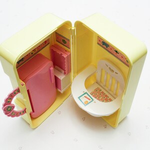 May include: A yellow plastic toy playset with a pink refrigerator, a white chair, and a white table. The playset is open and the interior is decorated with a floral pattern.