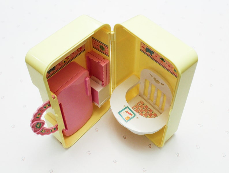 May include: A yellow plastic toy playset with a pink refrigerator, a white chair, and a white table. The playset is open and the interior is decorated with a floral pattern.