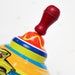 May include: A colorful spinning top with a red wooden handle. The top has a yellow, blue, and red design with a yellow car illustration.