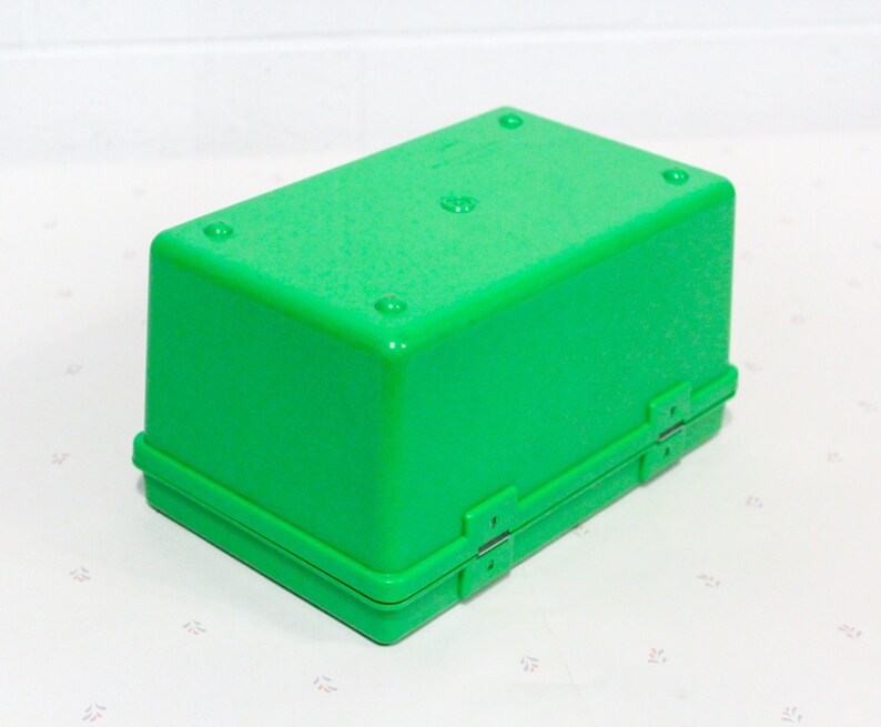 Puede incluir: Una caja de almacenamiento de pl&aacute;stico verde brillante con una tapa con bisagras y dos cierres. La caja est&aacute; cerrada y se encuentra sobre una superficie blanca.
