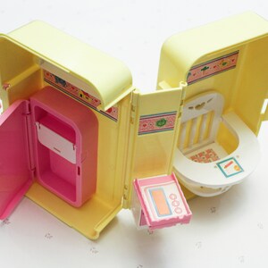 May include: A yellow and pink plastic toy playset with a kitchen and a nursery. The kitchen has a pink refrigerator with a door that opens. The nursery has a white high chair with a tray. The playset is designed for children to play pretend.
