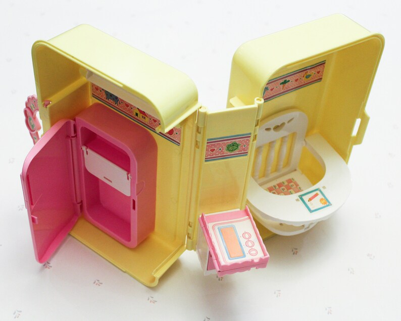 May include: A yellow and pink plastic toy playset with a kitchen and a nursery. The kitchen has a pink refrigerator with a door that opens. The nursery has a white high chair with a tray. The playset is designed for children to play pretend.