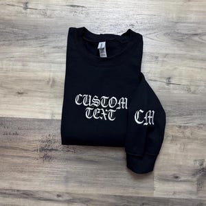 May include: Black sweatshirt with white embroidered text "CUSTOM TEXT" on the front and "CM" on the sleeve.