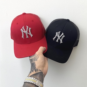 May include: A red and a black baseball cap with the New York Yankees logo in rhinestones. The red cap has a red brim and mesh back. The black cap has a black brim and mesh back. Both caps are held up against a white background.