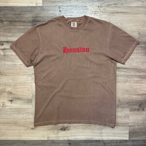 Puede incluir: Una camiseta marr&oacute;n con un bordado rojo de "Houston" en la parte delantera.