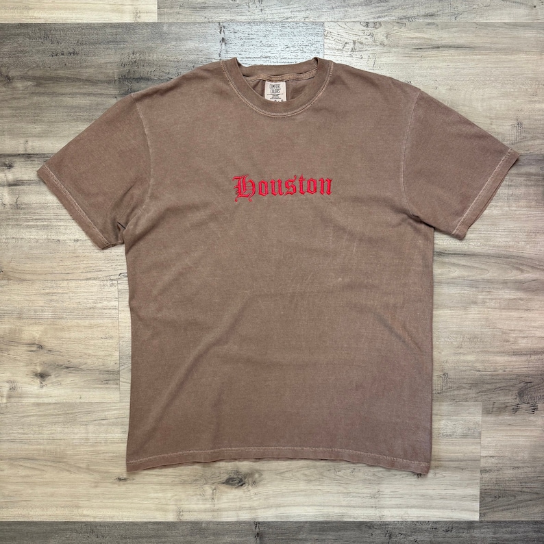 Puede incluir: Una camiseta marr&oacute;n con un bordado rojo de "Houston" en la parte delantera.