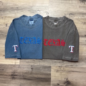 May include: Two vintage t-shirts with the word "TEXAS" embroidered on the front in blue and red. The shirts have a white "T" embroidered on the sleeves.