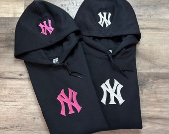 May include: Two black hooded sweatshirts with the New York Yankees logo embroidered on the front and hood. One logo is pink, the other is white. The sweatshirts have drawstrings and are displayed on a wooden surface.