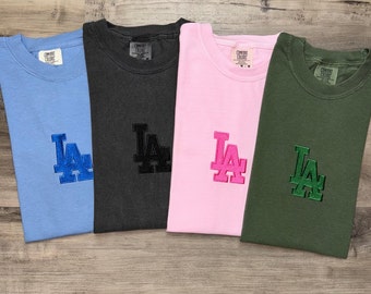 May include: Four t-shirts with the letters "LA" embroidered on the front. The shirts are in blue, black, pink, and green.