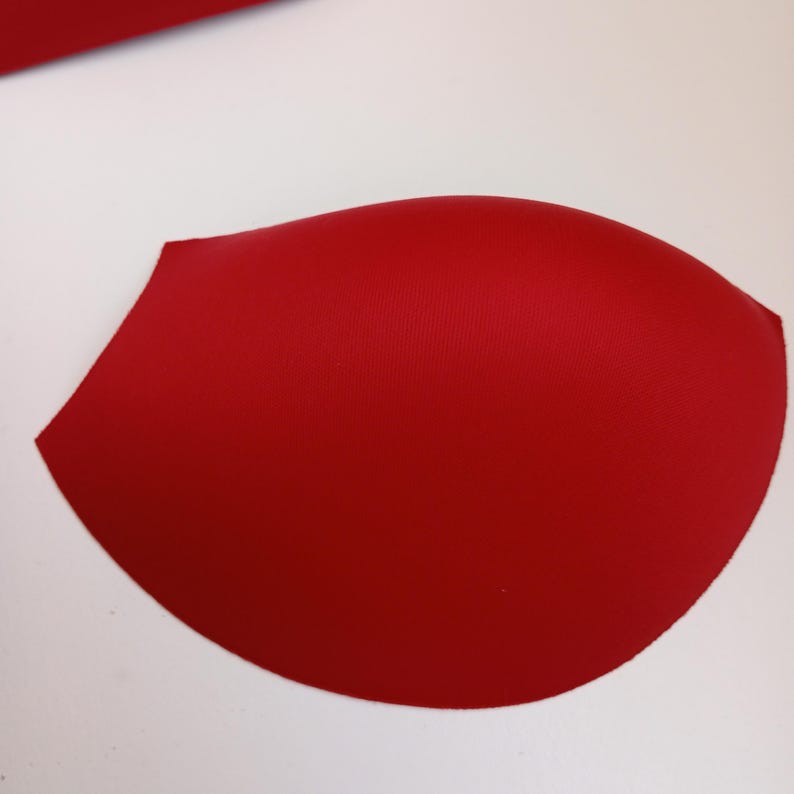 May include: A single, smooth, molded bra cup insert in a vibrant, solid red color. The cup features a rounded, contoured shape designed for garment support, with a clean, seamless surface and a curved edge profile on a plain white background.