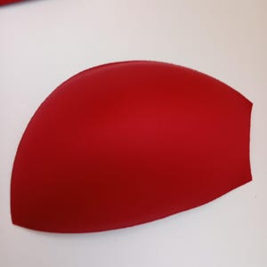 May include: A single, molded, semi-circular bra cup insert in a solid, vibrant red color. The item features a smooth, curved surface with a defined, rounded shape designed for garment padding, showcasing a consistent matte texture throughout.