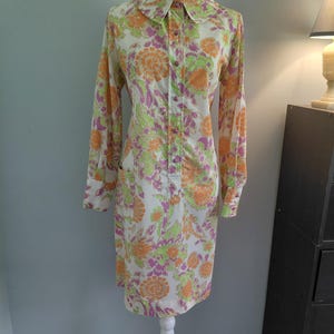 May include: A vintage-style dress with a floral pattern in orange, green, and purple on a white background. The dress has long sleeves, a collared neckline, and a button-down front. It is displayed on a white mannequin.
