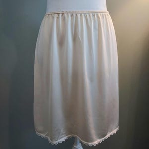 May include: Ivory-colored half slip with an elastic waistband and a delicate lace trim along the hem. The slip is displayed on a mannequin, showcasing its smooth, flowing fabric. A classic undergarment.