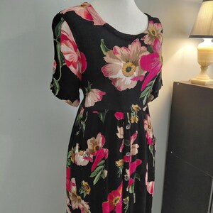 May include: A black midi dress featuring a vibrant floral pattern with large pink, peach, and green blossoms. The dress has short sleeves, a round neckline, and a vertical row of buttons running down the front center of the skirt portion.