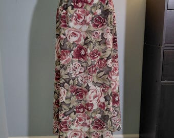 May include: A long floral skirt with a pattern of roses in shades of burgundy, pink, and cream. The skirt is displayed on a white mannequin, showcasing its full length and design. The background is a light blue wall.