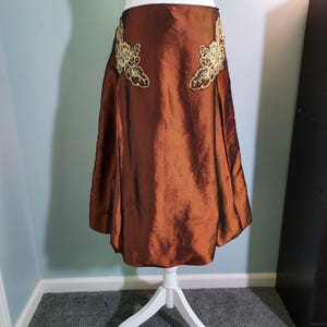 May include: A copper-colored skirt with a subtle sheen, displayed on a white mannequin. The skirt features intricate gold floral embroidery on both sides of the waist. The fabric drapes elegantly, creating a flowing silhouette. A stylish piece for special occasions.