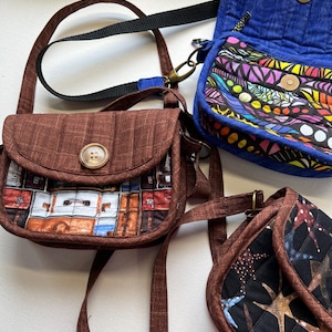 May include: Three small, handmade fabric purses in shades of brown and blue. One purse features a button closure and a print of vintage luggage. Another is blue with a quilted texture. The third has a black background with a starfish pattern.