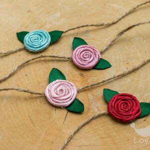 Könnte beinhalten: Vier Stoffrosen-Verzierungen mit grünen Blättern, die an einer Schnur befestigt sind. Die Rosen sind in den Farben Blau, Rosa, Rosa und Rot.