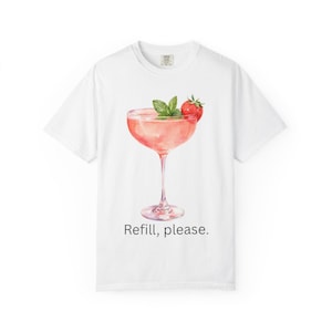 Puede incluir: Una camiseta blanca de manga corta con un gráfico de un cóctel rosa en una copa de tallo, adornado con una ramita de menta verde y una fresa roja. El texto "Refill, please." está impreso en negro debajo de la ilustración.