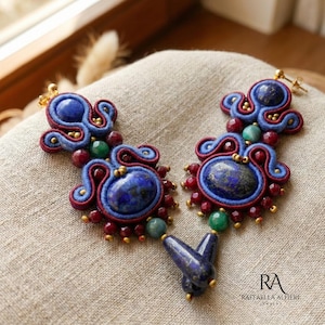 Può includere: Un paio di orecchini fatti a mano con grandi pietre di lapislazzuli blu, ovali e a goccia. Gli orecchini sono adornati con perline rosse e verdi e ricami blu e rossi. Gli accenti metallici color oro completano il design.