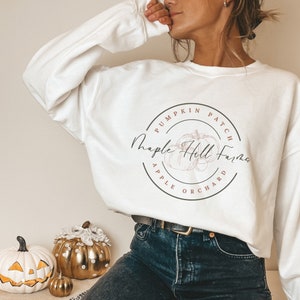 Puede incluir: Una sudadera blanca de manga larga con un logotipo circular que dice "Pumpkin Patch Maple Hill Farms Apple Orchard". El logotipo incluye una ilustración de calabaza. La sudadera se combina con vaqueros azul oscuro.