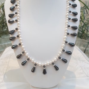 Puede incluir: Un collar de perlas blancas con cuentas en forma de lágrima, de color gris oscuro. El collar se exhibe sobre un busto blanco, mostrando el diseño elegante y los colores contrastantes. Una joya clásica.