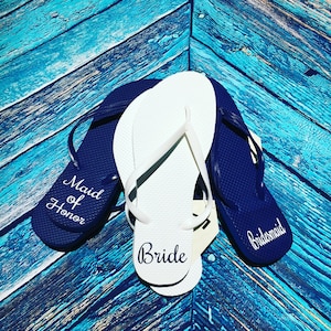 May include: Three pairs of flip-flops with the words "Bride", "Maid of Honor", and "Bridesmaid" printed on them. The flip-flops are white and navy blue.