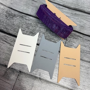 May include: A roll of purple ribbon with a textured finish, displayed on a tan cardboard spool holder. Three cat-shaped cardstock tags in white, gray, and tan are arranged below the ribbon, each with two horizontal slits.