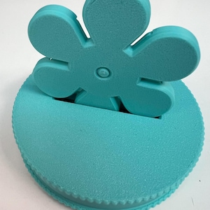 May include: A turquoise flower-shaped bottle stopper. The stopper has a textured surface and a round base with a scalloped edge. The flower has five petals and a small circular detail in the center.