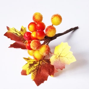 May include: A small artificial autumn floral arrangement with yellow and orange berries and brown and yellow leaves.