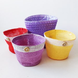 Op de afbeelding: Vier kleine gevlochten mandjes in rood, geel en twee tinten paars. Elk mandje heeft een witte band rond de bovenrand en een enkele lichtgekleurde houten knoop in het midden aan de voorzijde. De mandjes hebben een textuur met open weving.