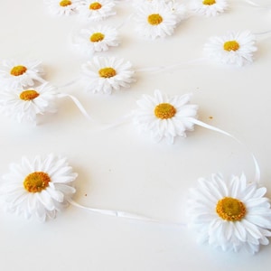Puede incluir: Una guirnalda de margaritas blancas con centros amarillos. La guirnalda está enhebrada en una cinta blanca y es perfecta para agregar un toque de primavera a cualquier espacio.