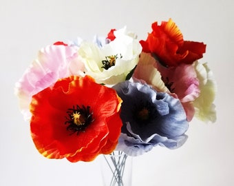 May include: A bouquet of artificial poppy flowers in vibrant shades of red, blue, white, and soft pink, featuring delicate ruffled petals and dark central stamens. The flowers are arranged in a clear glass vase with thin, visible wire stems.